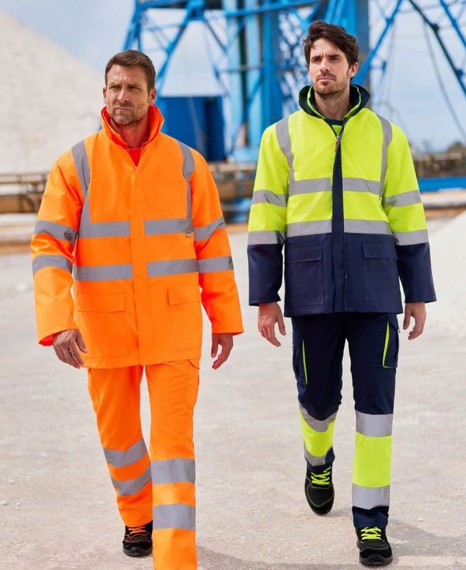 Dos operarios caminando por una cantera con ropa de alta visibilidad: uno con conjunto naranja flúor de chaqueta y pantalón con bandas retrorreflectantes, otro con chaqueta bicolor amarillo flúor y azul marino y pantalón a juego.